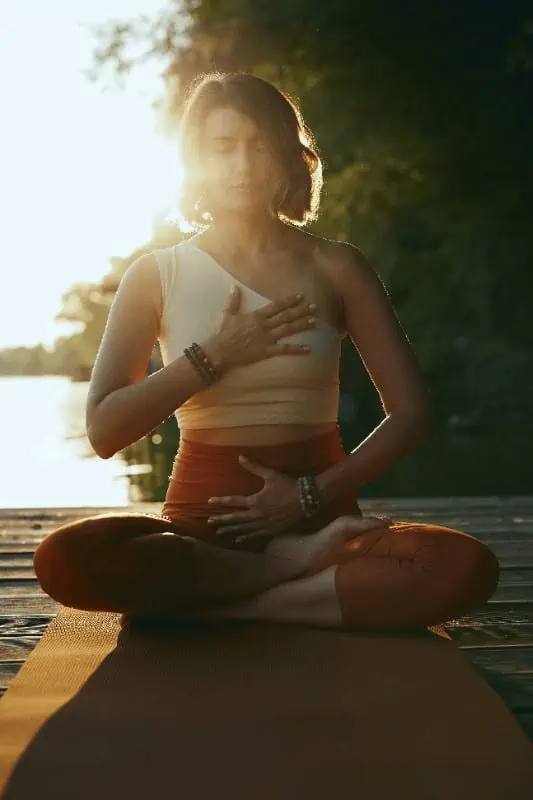 A woman sits meditating with eyes closed, her hands placed on her abdomen and chest. The sun shines behind her, illuminating.