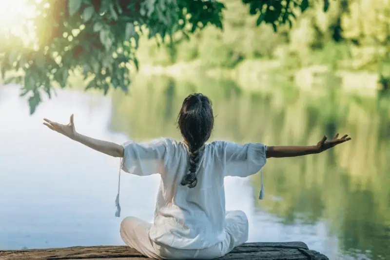 A woman sits in front of a lake in a meditative position, arms outstretched to receive the energies of yoga