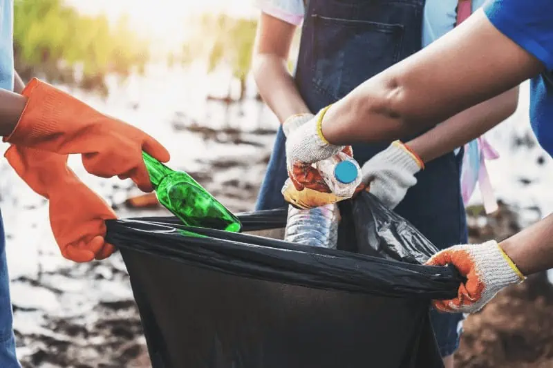 A group of people hold a large trash bag, filling it with bottles and trash. Community service is an example of karma yoga.