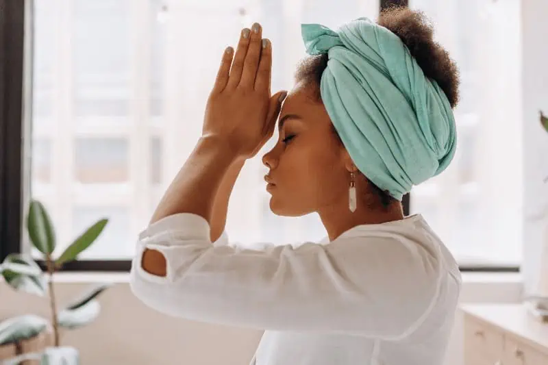 A woman meditates with her hands in a prayer position (anjali mudra) placed at her third eye in the center of her forehead.
