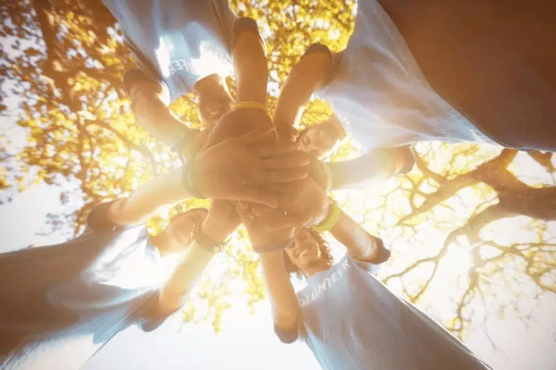 A group of volunteers stand in a circle, placing their hands together, showing they are coming together in an act of service.