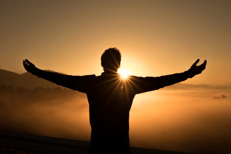 A silhouetted person stands on top of a mountain, arms outstretched, facing the sun, signifying the feeling of spiritual liberation
