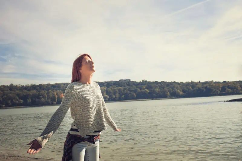 A woman stands in front of a tranquil lake, arms outstretched, with a peaceful demeanor reflecting the effects of karma yoga.