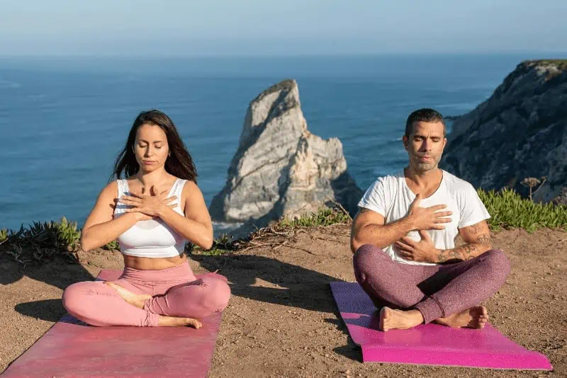 man and woman sitting on their yoga mats, practicing breathing techniques with a beautiful ocean view background