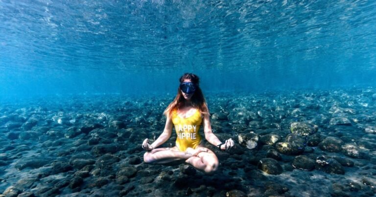 The image shows a person underwater, sitting cross-legged in a meditative pose. The individual is wearing a yellow swimsuit with the text "HAPPY HIPPIE" printed on it. The underwater setting is clear, with a rocky seabed visible below. The person appears to be floating slightly above the seabed, creating a serene and surreal atmosphere. This scene captures a moment of calm and mindfulness in a unique underwater environment.