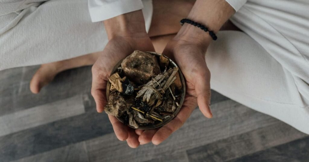 The image shows a person seated cross-legged on the floor, dressed in white clothing, holding a small bowl filled with natural elements like dried herbs, leaves, and stones or roots. A black beaded bracelet is visible on their wrist, adding detail to the serene scene. The image evokes a connection to Ayurveda, emphasizing the use of natural remedies to support joint health and overall wellness. Combined with yoga practices, this approach offers holistic benefits for physical and mental well-being.