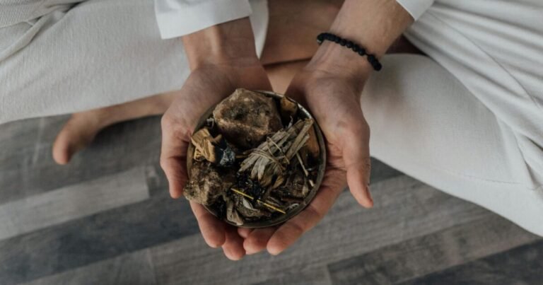 The image shows a person seated cross-legged on the floor, dressed in white clothing, holding a small bowl filled with natural elements like dried herbs, leaves, and stones or roots. A black beaded bracelet is visible on their wrist, adding detail to the serene scene. The image evokes a connection to Ayurveda, emphasizing the use of natural remedies to support joint health and overall wellness. Combined with yoga practices, this approach offers holistic benefits for physical and mental well-being.