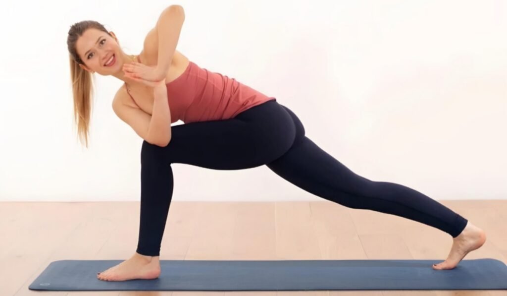 A person is performing a yoga pose on a blue yoga mat. They are in a lunge position with the right leg bent at a 90-degree angle and the left leg extended straight back. Their torso is twisted to the right, with their hands pressed together in a prayer position near the chest. They are wearing a pink tank top and black leggings. The background is plain white.