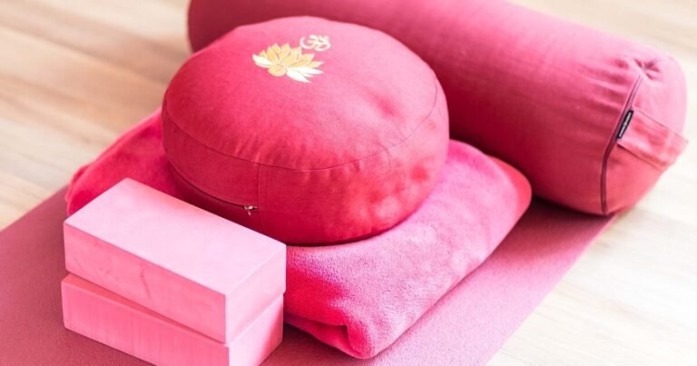The image shows a collection of yoga and meditation accessories placed on a wooden floor. There is a round red cushion with a lotus flower and Om symbol embroidered on it, a cylindrical bolster pillow, two pink yoga blocks, a folded pink blanket, and a pink yoga mat. The items are neatly arranged, suggesting a setup for a comfortable and supportive yoga or meditation session.