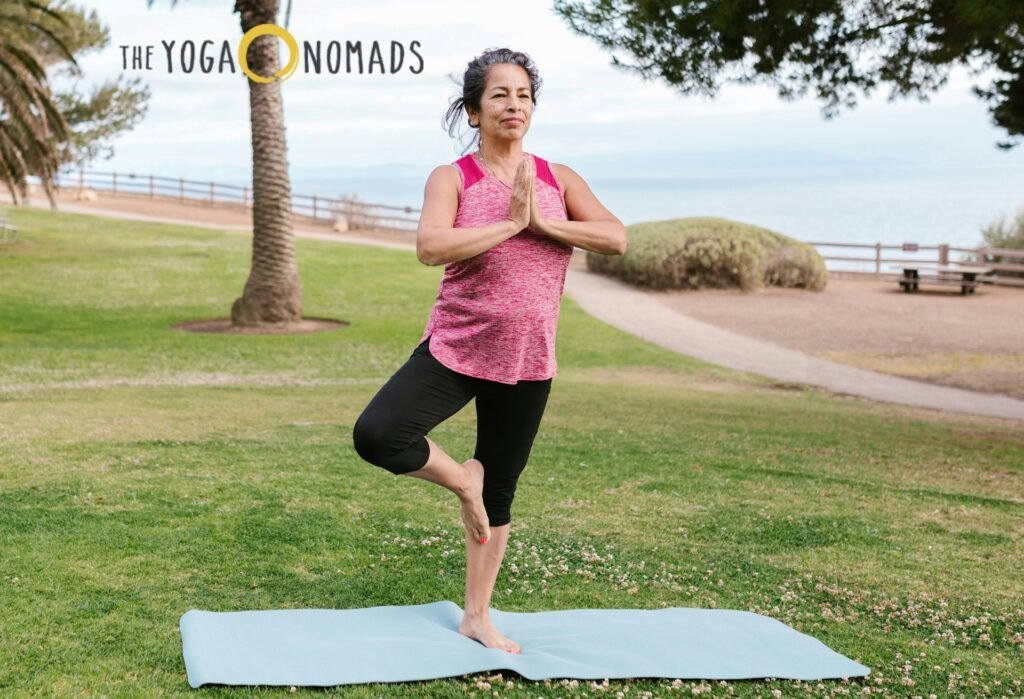 Comprehensive Guide to Yoga for Joint Pain yoga for joint pain 5 A person practices the tree pose on a blue yoga mat in an outdoor setting with a scenic ocean view. Standing on one leg, they press their hands together in a prayer position at chest level, promoting balance and joint stability. The background features lush greenery, a palm tree, and a pathway leading to the shore. The text 'THE YOGA NOMADS' appears in the top left corner of the image. This calming environment emphasizes yoga’s benefits for joint health, strength, and mobility.