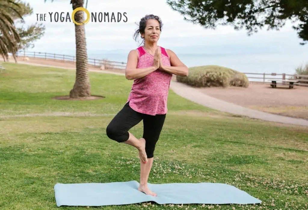 A person practices the tree pose on a blue yoga mat in an outdoor setting with a scenic ocean view. Standing on one leg, they press their hands together in a prayer position at chest level, promoting balance and joint stability. The background features lush greenery, a palm tree, and a pathway leading to the shore. The text &#039;THE YOGA NOMADS&#039; appears in the top left corner of the image. This calming environment emphasizes yoga’s benefits for joint health, strength, and mobility.