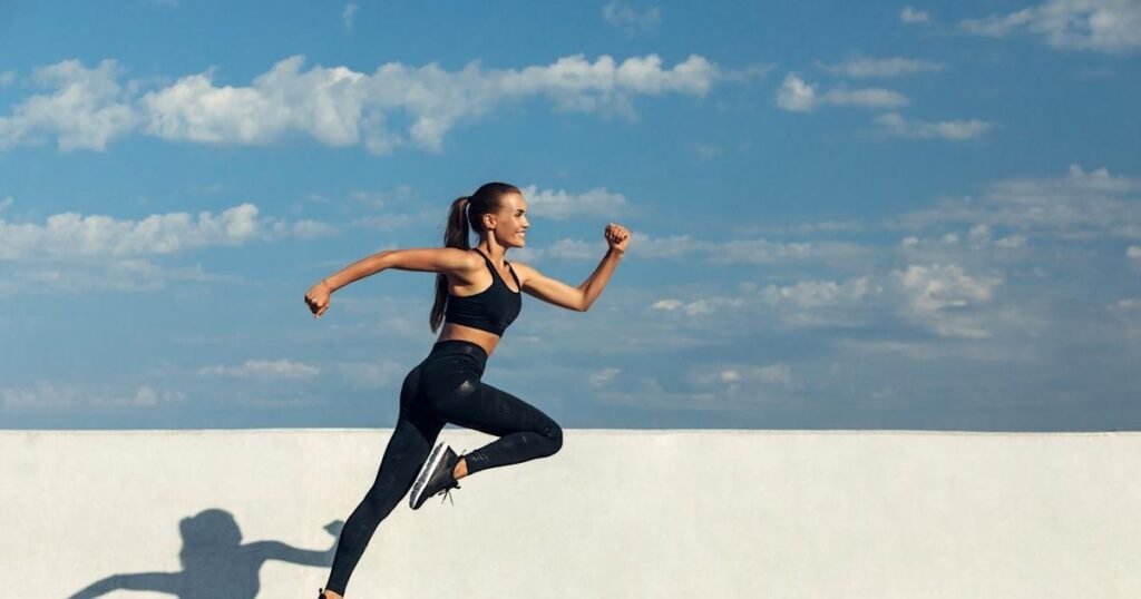 A person leaps mid-air in a powerful running pose against a bright blue sky scattered with clouds. They wear a black sports bra, black leggings, and supportive black running shoes—captured above a white wall casting a distinct shadow on the ground below. The dynamic stance and choice of footwear emphasize impact protection and joint support, relevant to discussions about ideal shoes for reducing yoga-related joint pain.