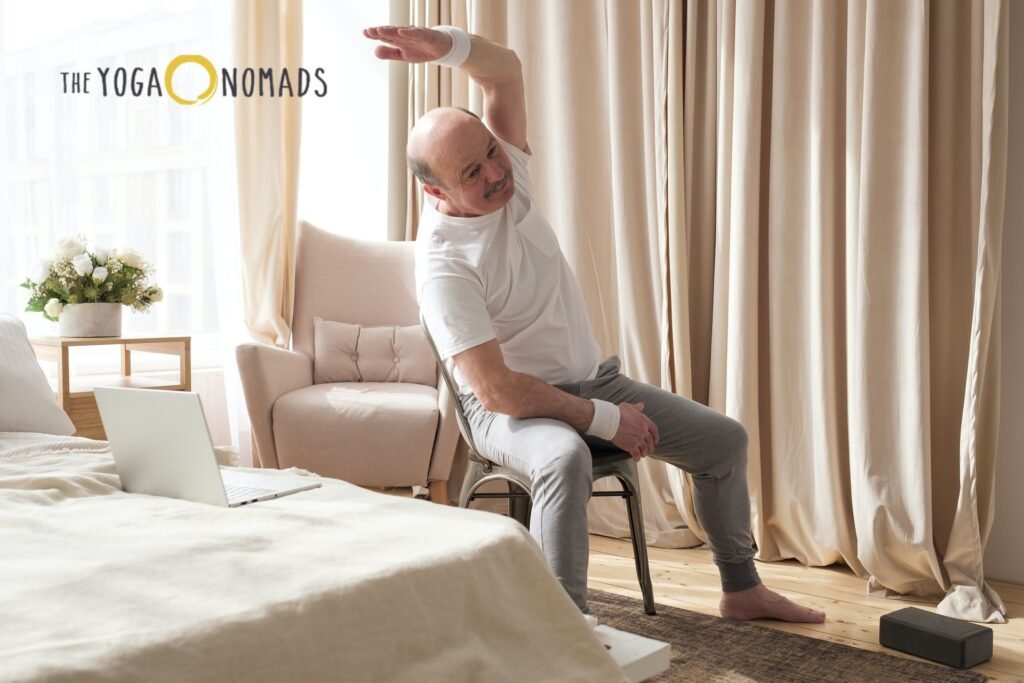 5 Top-Rated Online Yoga Classes for Joint Pain Management online yoga classes for joint pain 3 A person seated on a chair performs a gentle side stretch, raising one arm overhead while the other rests on their leg. Dressed in a white t-shirt and gray pants, they sit in a softly lit room with beige curtains and a cozy armchair nearby. A yoga block is placed on the wooden floor, and a laptop on the bed in front suggests participation in an online yoga class. The scene is calm and accessible, underscoring how virtual sessions can accommodate joint-friendly movement in everyday spaces.