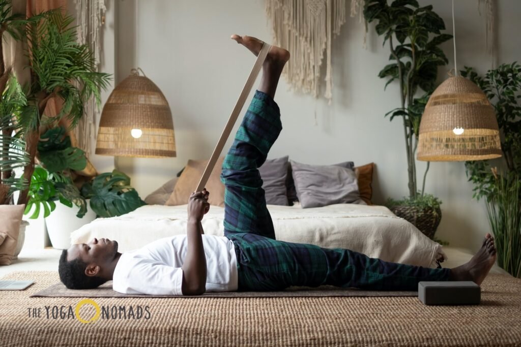 5 Top-Rated Online Yoga Classes for Joint Pain Management online yoga classes for joint pain 7 A person lies on a yoga mat in a softly lit, bohemian-style room, using a yoga strap to gently hold one leg extended upward while keeping the other leg grounded. They wear a white top and plaid pants, demonstrating a supportive stretch for joint flexibility. Surrounding the scene are cozy furnishings—including a bed with cushions, houseplants, woven lamps, and a macramé wall hanging—evoking a tranquil home environment. The image underscores the accessibility and therapeutic benefits of online yoga for joint health in everyday spaces.