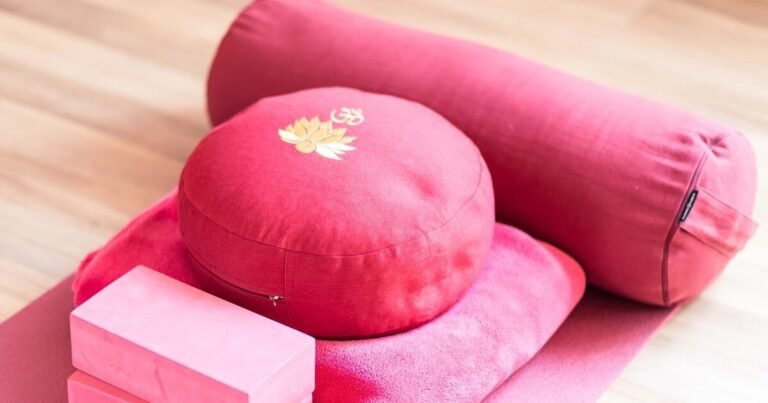 A collection of yoga props arranged on a wooden floor, including a red cylindrical bolster, a round red cushion embroidered with a lotus and Om symbol, two stacked pink yoga blocks, and a folded pink blanket. These supportive props are commonly used to enhance comfort and alignment during yoga practice, especially for individuals seeking joint-friendly modifications.