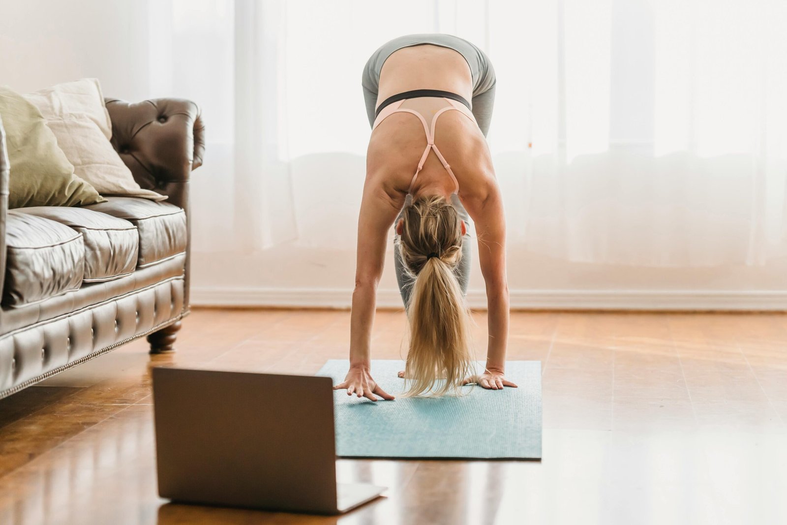 A person practices yoga indoors on a blue mat, performing a forward bend with hands touching the mat and head hanging down. They wear a light pink sports bra and gray leggings, facing away from the camera. An open laptop is placed in front of the mat, suggesting participation in an online yoga session. The setting resembles a cozy living room, with a brown leather couch, cushions, sheer white curtains, and natural light filtering in. The image evokes a calm, supportive environment for home-based yoga, ideal for gentle movement and joint-friendly routines.