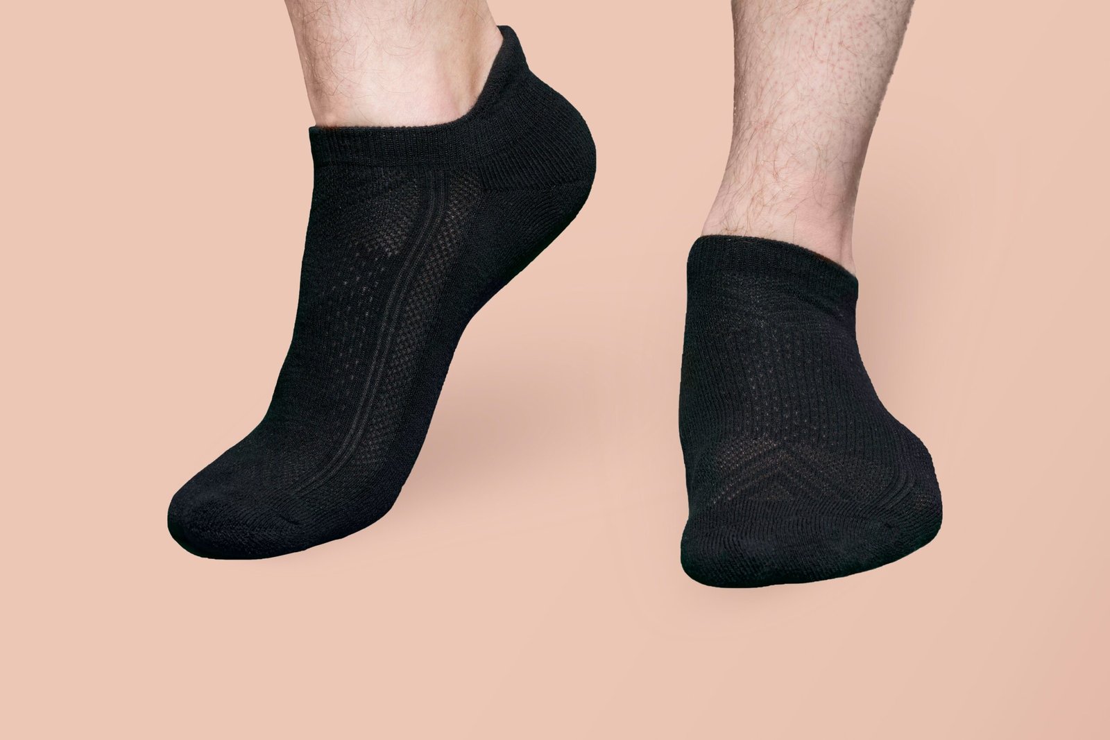 A pair of feet wearing black ankle-length yoga socks is shown against a plain beige background. The socks are low-cut with a subtle textured pattern, covering the feet just up to the ankles. The feet are slightly lifted off the ground, suggesting motion or a poised yoga stance. The image emphasizes the minimalist design and snug fit of anti-slip yoga socks, ideal for pain-free practice and enhanced stability during movement.